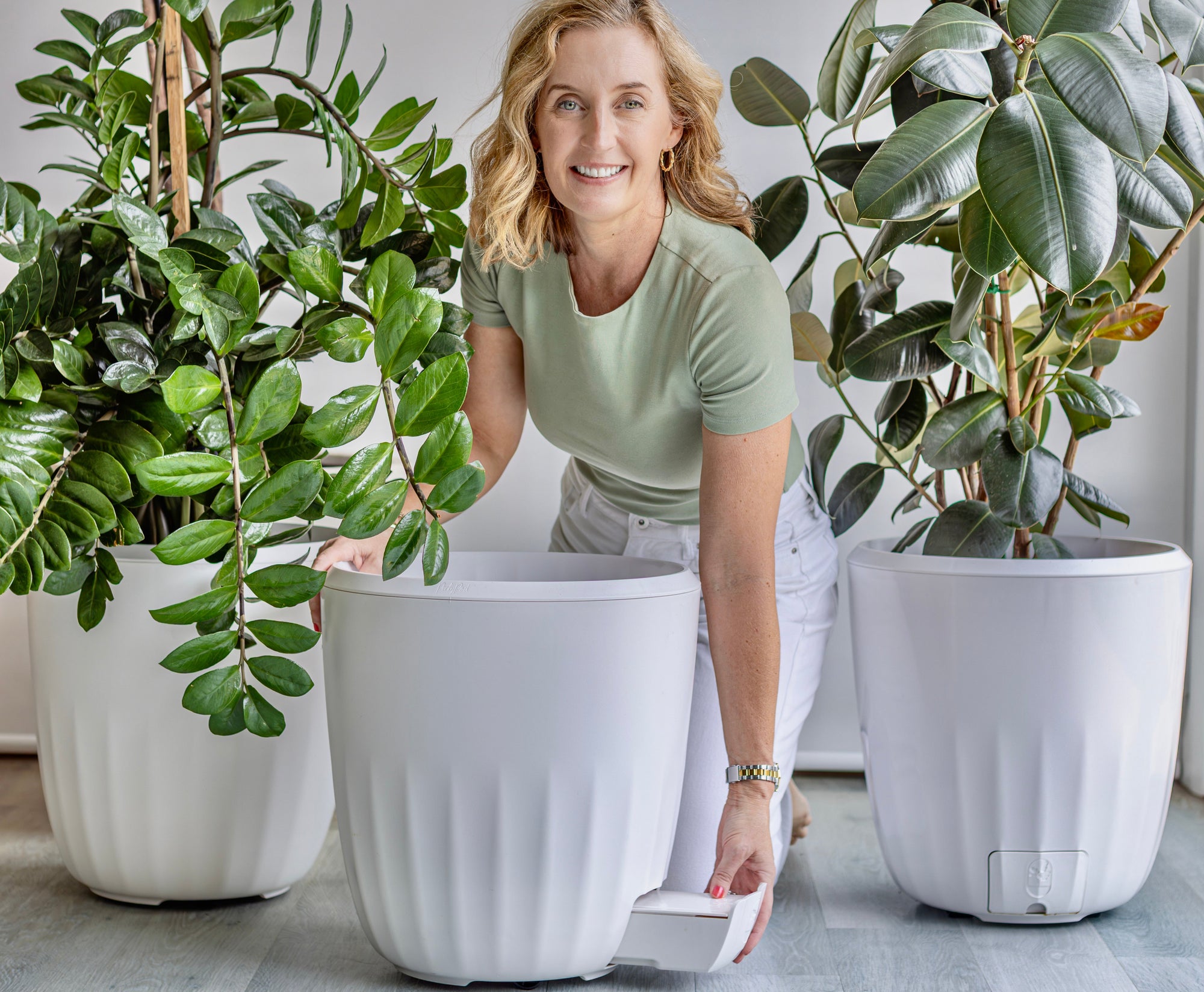 Louise is wearing a green tshirt and is bent over a large white PerkyPod plant pot while opening the water catchment drawer. She is flanked by two large, lush plants which are also in PerkyPod plant pots.