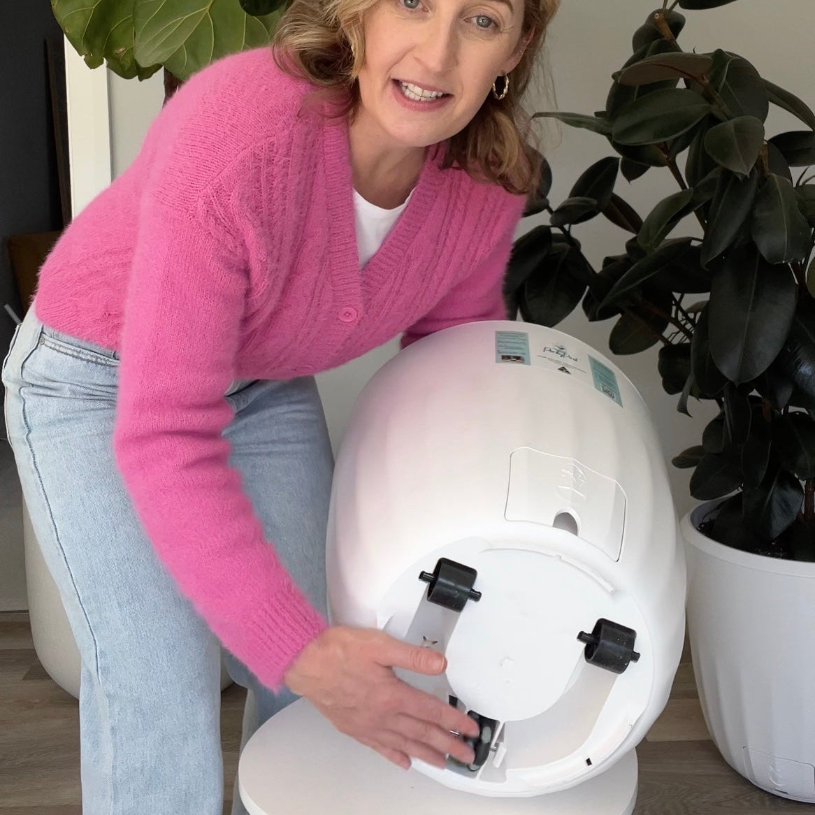 Woman in pink cardigan holds PerkyPod sideways to show the hidden wheel system underneath. Lush, large plants are in the background